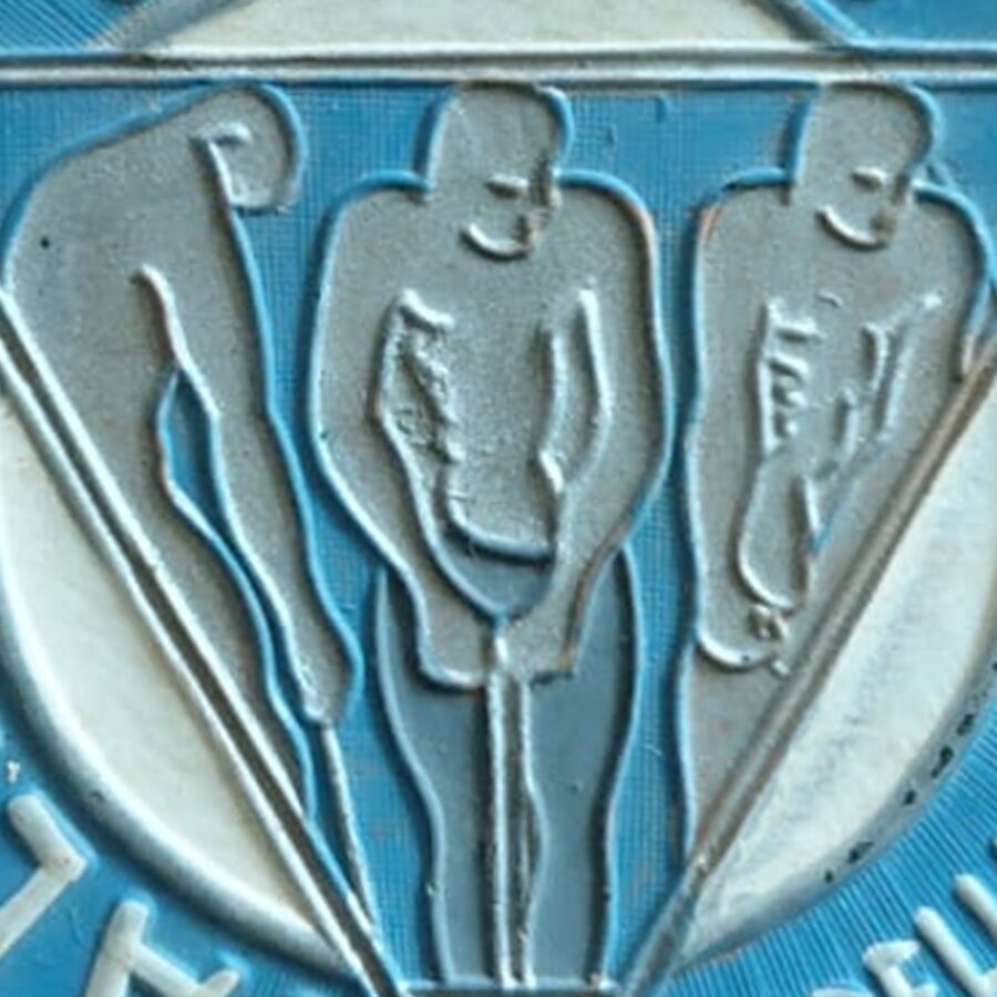 Detalle del medallón: tres figuras de pie dentro de un triángulo invertido, en relieve sobre esmalte azul