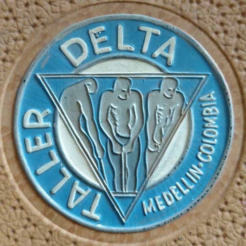 Medallón original del Taller Delta: placa esmaltada en azul con un triángulo invertido y tres figuras, con la inscripción Taller Delta, Medellín, Colombia